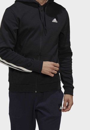 Mann trägt schwarzen Adidas Zip-Hoodie mit weißen Streifen an den Ärmeln und schwarze Adidas Hose mit Logopatch am Oberschenkel.