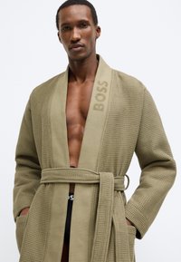 Accappatoio di colore verde oliva con trama a cialda, cintura in vita e la parola "BOSS" ricamata sul colletto. Tasche frontali incluse.
