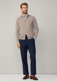 Cardigan zippé beige sur une chemise à col bleu clair, associé à un pantalon navy foncé et des chaussures de ville marron. Texture lisse et design ajusté.