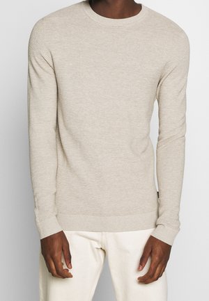 Homme portant un pull à manches longues beige clair côtelé et un pantalon blanc cassé, debout devant un fond uni.