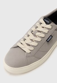 Light grey suede sneaker with cream-colored laces, featuring black brand tags. Smooth texture and rubber sole with a textured grip pattern.
