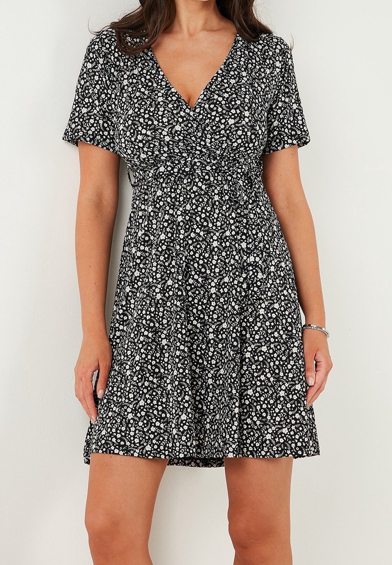 Femme portant une robe noire à manches courtes avec un motif floral blanc et un col en V, debout contre un mur blanc uni.