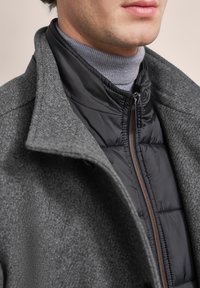 Grauer Wollmantel mit großem Kragen über einer schwarzen Daunenjacke. Die Daunenjacke verfügt über einen Reißverschluss und sichtbare Steppung, darunter ist ein grauer Rollkragenpulli.