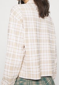 Béžov�á kostkovaná bavlněná košile s bílými pruhy, dlouhými rukávy a volnějším střihem, s zaobleným spodním lemem bez viditelných zapínání nebo ozdob.