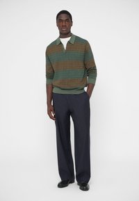 Adīta džemperis ar zaļām un brūnām svītrām, ar apkakli un tekstūru. Kombinēts ar tumši ziliem biksēm ar smalkām svītrām un melniem apaviem; modelis nostājies pozā.