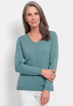 Femme aux longs cheveux gris portant un pull en V bleu sarcelle et un pantalon blanc, debout devant un fond clair.