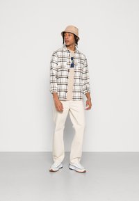 Camisa de franela de cuadros en blanco, marrón y negro sobre una camiseta beige, combinada con pantalones color crema y zapatillas blancas. Sombrero de cubo beige.