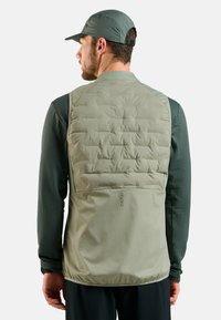 Mann in gepolsterter olivgrüner Weste und passender Kappe, von hinten blickend, mit strukturiertem Langarmshirt und dunkler Hose vor weißem Hintergrund.