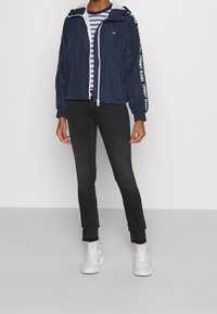 Person wearing navy zip-up jacket with white "Tommy Jeans" text on sleeves, striped shirt, black jeans, and white sneakers, standing upright.