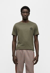 Χακί βαμβακερό T-shirt με κοντά μανίκια, με μικρό μαύρο σχέδιο κάκτου στο στήθος. Φορεμένο με καρό παντελόνι με σχέδια.