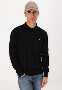 Svart långärmad polo T-shirt med krage och tre knappar, prydd med en liten gul fågel-logotyp på vänster bröst. Kombineras med blå jeans.