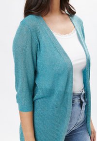 Cardigan de punto color teal con un corte holgado, mangas tres cuartos y escote en V, que se lleva sobre una blusa blanca y unos vaqueros azules.
