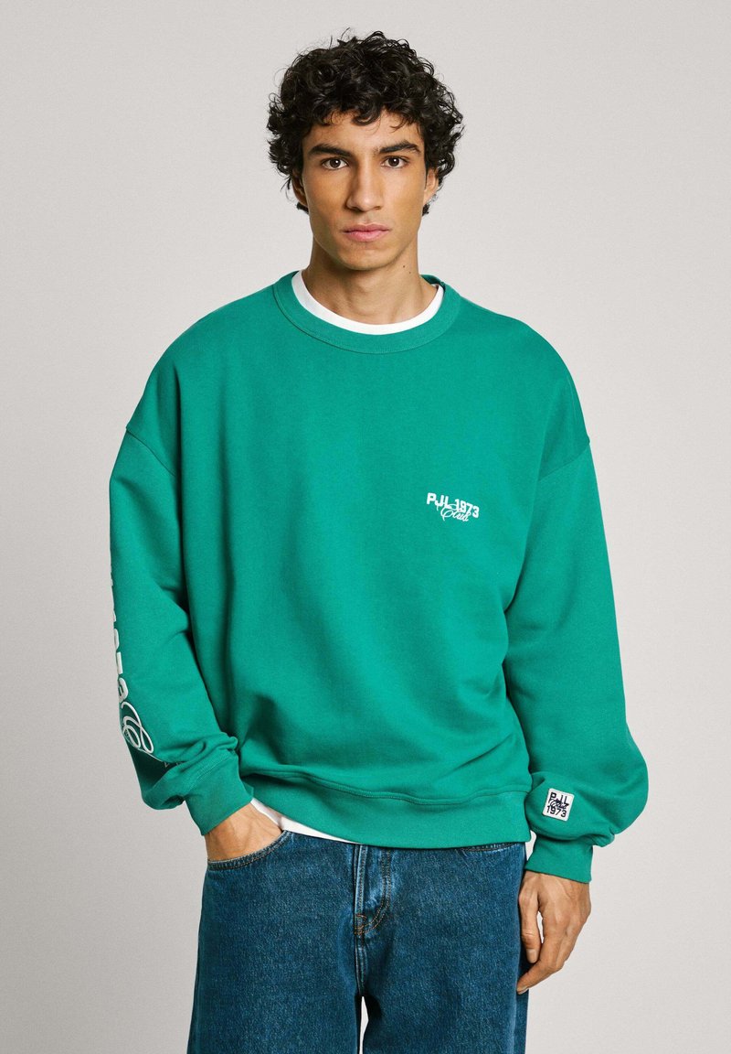 Groene sweatshirt met een ontspannen pasvorm, ronde halslijn en wit logo op de borst. Voorzien van lange mouwen en ribgebreide manchetten. Gegdraagt met blauwe jeans.