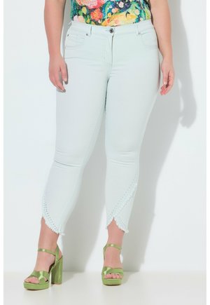 Kvinde iført hvide slim-fit jeans med bølgede kanter og hulmønster detaljer, kombineret med grønne højhælede sandaler og en blomster top.