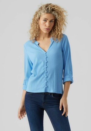 Camicia azzurra chiara, con texture, a maniche lunghe con collo a V e maniche arrotolate, abbinata a jeans scuri. Presenta dettagli di bottoni sulla parte anteriore.