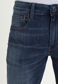 Dark blue denim jeans with a classic five-pocket design. Features visible stitching, a zipper fly, and a metal button at the waistband.