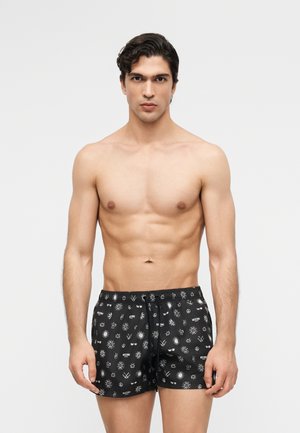 Muž bez trička s krátkymi tmavými vlasmi, oblečený v čiernych plavkových šortkách s bielymi grafickými symbolmi a logami na jednoduchom svetlom pozadí.