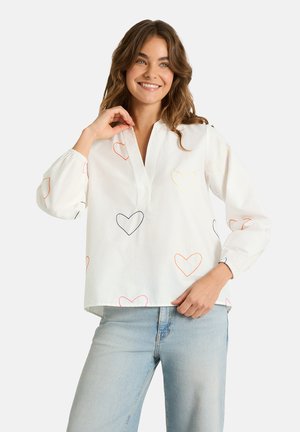 Lächelnde Frau in weißer langärmeliger Bluse mit mehrfarbigen, bestickten Herzen und hellblauer Jeans vor einfachem Hintergrund.