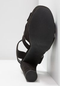 Escarpin en daim noir à talon haut avec des brides croisées autour de la cheville et une semelle en caoutchouc texturée, vu de côté pour mettre en évidence le talon et le design.