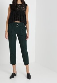 Černý krajkový crop top s volánkovými rukávy, spárovaný s tmavozelenými řasenými kalhotami se širokým pasem a zúženým střihem na kotníku. Černé muly.