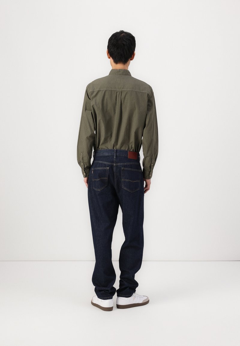 Olivgrünes Hemd mit Knopfleiste, lässige dunkelblaue Jeans mit Stickdetails, braunes Lederlabel am Bund, weiße Sneakers.