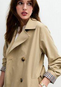 Beige double-breasted trenchcoat met grote bruine knopen, notch lapel en geruite manchetten. Zacht, glad materiaal met een ontspannen pasvorm.