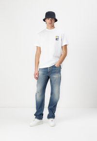 Hvit t-skjorte med grafikk foran, lyseblå ripped jeans og svart bøttehatt. Modellen har på seg hvite sneakers med teksturert såle.