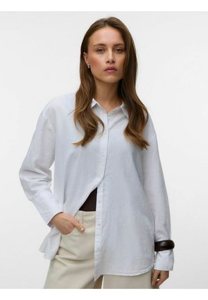 Frau mit langen braunen Haaren trägt ein lockeres weißes Hemd über beigen Hosen, Hand in der Tasche und ein dunkles Armband am linken Handgelenk.