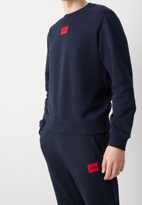 Felpa blu navy con polsini e orlo a costine, con un logo rettangolare rosso sul petto. Texture liscia, design informale.