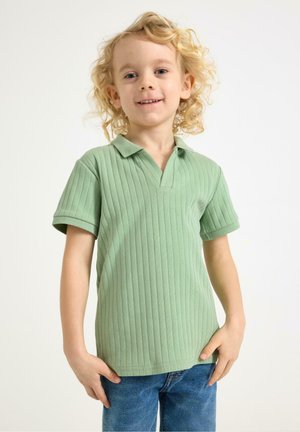 Ungt barn med krøllet blondt hår iført en grøn ribstrikket kortærmet skjorte og blå jeans, stående mod en hvid baggrund.