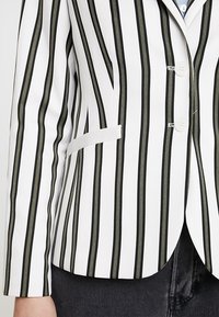 Proužkované sako v bílé barvě s černými a olivově zelenými pruhy. Disponuje zapínáním na jeden knoflík a přední kapsou. Hladká texturou látky.