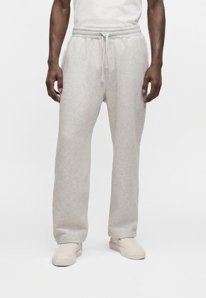 Homme portant un pantalon de survêtement gris clair avec cordon de serrage et des chaussures à enfiler écrues sur un fond blanc uni.