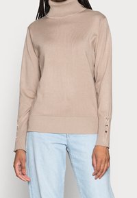 Pull à col roulé beige en maille douce, avec des poignets côtelés ornés de boutons décoratifs et une coupe ajustée. Assorti à un jean bleu clair.
