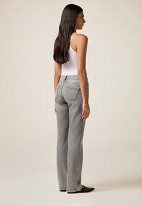 Pantalones de mezclilla gris de pierna ancha con acentos de botones laterales, diseñados con un corte de tiro medio. Combinados con una camiseta sin mangas blanca, modelo visto desde atrás.