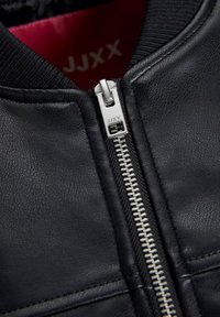 Primo piano di una cerniera in argento su una giacca di pelle nera con "JJXX" incisa sul tirazip e foderatura interna rossa visibile.