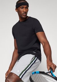 Μαύρο κοντομάνικο μπλουζάκι συνδυασμένο με γκρίζες αθλητικές βερμούδες που έχουν πράσινες και μπλε ρίγες. Το μοντέλο κρατάει μια ρακέτα τένις και φοράει ένα μαύρο κορδέλα για το κεφάλι.