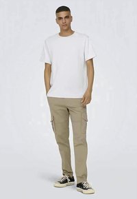 Ung mand står med den ene hånd i lommen, iført hvid t-shirt, beige cargo-bukser og sorte sneakers med lave snuder og hvide snørebånd.