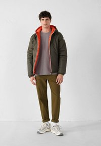 Giacca trapuntata verde con fodera arancione, chiusura con cerniera e cappuccio. Indossata sopra una maglietta grigia e abbinata a pantaloni oliva e sneakers.