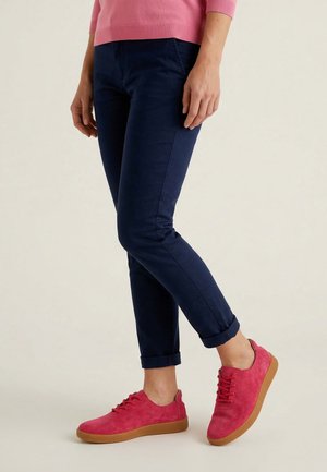 Persoon die marineblauwe omslagbroek, roze trui en felroze veterschoenen met bruine zolen draagt, staand op een lichte vloer.