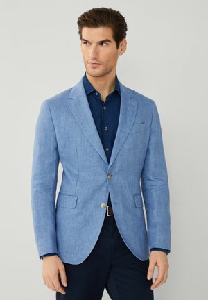 Blazer de lino azul claro con solapa de muesca, dos bolsillos frontales y cierre de un solo botón, usado sobre una camisa azul marino.