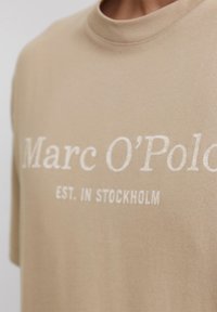 Béžové tričko vyrobené z jemnej bavlny, so zaobleným výstrihom a vo voľnom strihu. Na prednej strane je biely nápis: "Marc O'Polo EST. V STOCKHOLME."