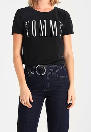 Vrouw in een zwart T-shirt met de tekst "TOMMY" en donkerblauwe high-waisted spijkerbroek met een ronde zilveren riemgesp.