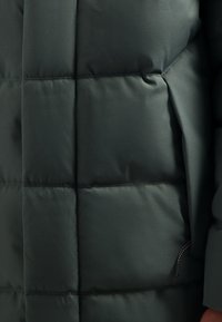 Donkergroene pufferjack, gemaakt van een gladde stof, met een doorgestikt patroon, een zijzak met rits en een minimalistisch ontwerp.