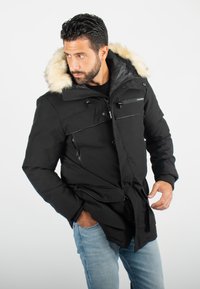 Parka noire avec capuche en fausse fourrure, poches zippées, cordons de serrage ajustables et coupe décontractée, porté sur un jean bleu clair.
