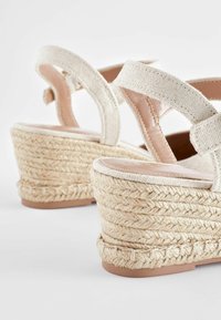 Sandales compensées beiges avec semelles compensées en jute tressé et brides de cheville en tissu sur fond blanc.