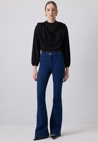 Top lungo a maniche lunghe nero drappeggiato abbinato a jeans a zampa d'elefante blu a vita alta. I jeans presentano un classico design a cinque tasche e cuciture sul retro.