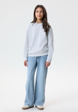 Noor tüdruk seisab, seljas helehall dressipluus, laiad helerohelised teksad ja valged tossud, taustaks üksluine valge sein.