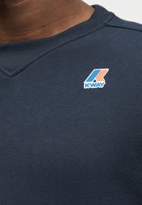 Σκούρο μπλε φούτερ με μικρό λογότυπο K-Way που περιλαμβάνει μπλε και πορτοκαλί ρίγες στο πάνω αριστερό μέρος του στήθους.