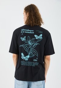 Sorte oversized t-shirt med et blåt grafik af sommerfugle og en lilje, samt teksten "Overflod er et tankesæt" og året "1994."