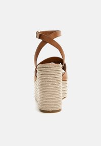 Hnědé kožené sandály na podpatku s křížovým páskovým designem. Mají texturovanou, tkanou krémovou rope platformu a robustní blokový podpatek.